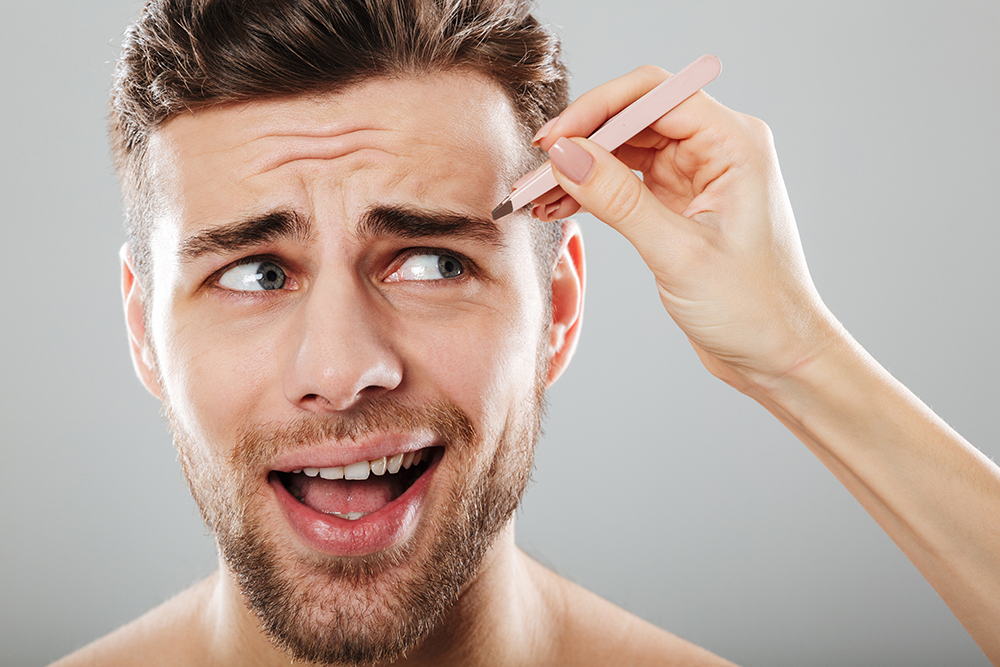 Female hand plucking scared men's eyebrows with tweezers isolated over gray background